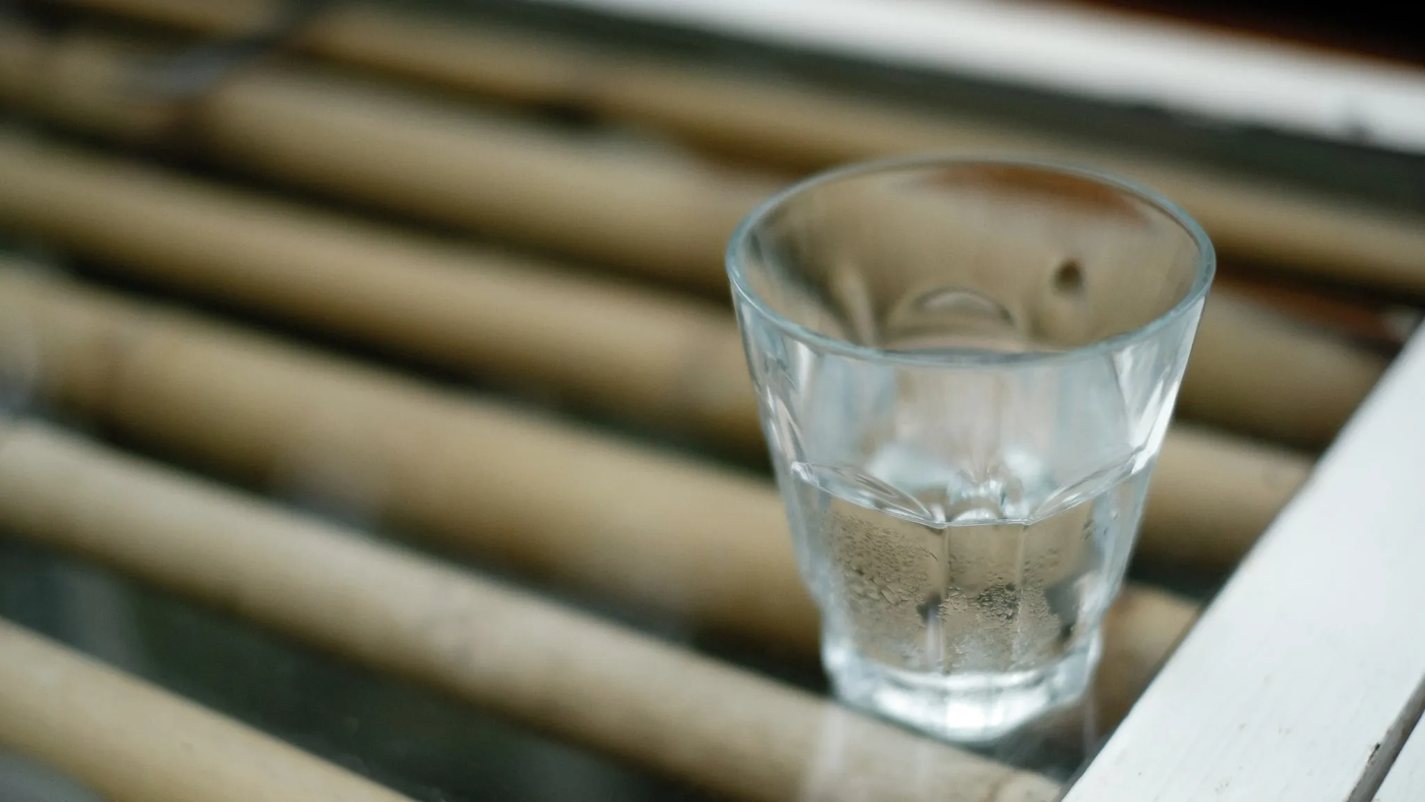 Een nachtkastje met een eenvoudig glas water, ochtendlicht door een dun gordijn.